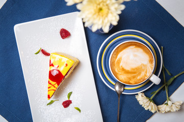 Wall Mural - Top view on sweet dessert piece of cake with yellow icing and red sauce with a cup of cappuccino coffee on the blue napkin on the table decorated with flower, horizontal format