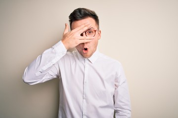 Sticker - Young handsome business mas wearing glasses and elegant shirt over isolated background peeking in shock covering face and eyes with hand, looking through fingers with embarrassed expression.