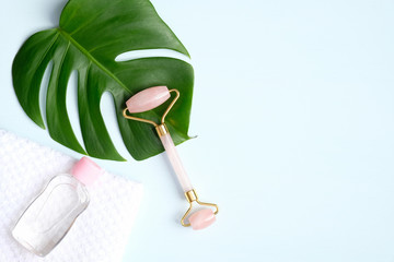 Wall Mural - Rose quartz face roller and essential oil with monstera leaf on blue background. Flat lay, top view. Massage tool for facial skin care, SPA beauty treatment concept