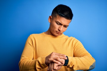 Young handsome latin man wearing yellow casual sweater over isolated blue background Checking the time on wrist watch, relaxed and confident