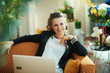 © Alliance - smiling elegant female writing on laptop while sitting on couch