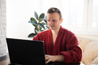 © vitalii_demin - A young man smiles as he reads on a laptop screen while relaxing on a comfortable sofa at home in dressing gowns