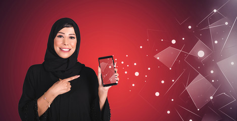 Arab happy beautiful young woman with hijab holding blank screen mobile phone and pointing finger over red background. Arab technology concept.
