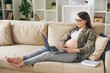 © pressmaster - Attractive young pregnant woman in homewear sitting on sofa and reading article on laptop while making notes about maternity