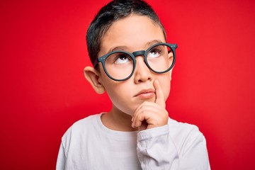 Wall Mural - Young little smart boy kid wearing nerd glasses over red isolated background serious face thinking about question, very confused idea