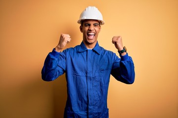 Poster - Young handsome african american worker man wearing blue uniform and security helmet celebrating surprised and amazed for success with arms raised and open eyes. Winner concept.