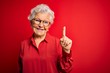 Senior beautiful grey-haired woman wearing casual shirt and glasses over red background showing and pointing up with finger number one while smiling confident and happy.