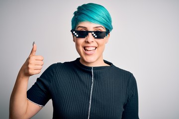 Sticker - Young woman with blue fashion hair wearing thug life sunglasses over white background doing happy thumbs up gesture with hand. Approving expression looking at the camera showing success.