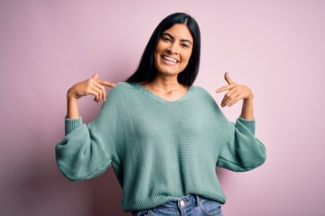 Young beautiful hispanic fashion woman wearing green sweater over pink background looking confident with smile on face, pointing oneself with fingers proud and happy.