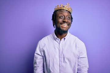 Wall Mural - Young african american man wearing golden crown of king over isolated purple background looking away to side with smile on face, natural expression. Laughing confident.