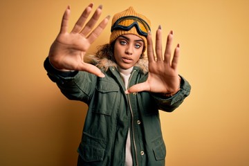 Wall Mural - Young african american afro skier girl wearing snow sportswear and ski goggles doing frame using hands palms and fingers, camera perspective