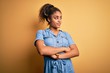 © Krakenimages.com - Young beautiful african american girl wearing denim dress standing over yellow background looking to the side with arms crossed convinced and confident