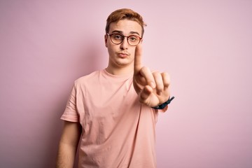 Young handsome redhead man wearing casual t-shirt standing over isolated pink background Pointing with finger up and angry expression, showing no gesture