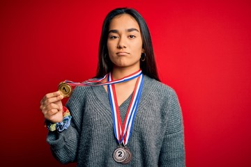 Young beautiful successful asian woman wearing medals standing over red background with a confident expression on smart face thinking serious