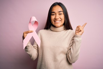 Canvas Print - Young beautiful asian woman holding pink cancer ribbon symbol over isolated background very happy pointing with hand and finger to the side