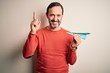 © Krakenimages.com - Middle age hoary man holding paper aircraft standing over isolated white background surprised with an idea or question pointing finger with happy face, number one