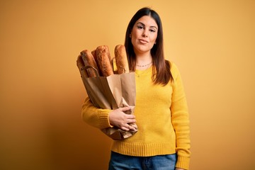 Canvas Print - Young beautiful woman holding a bag of fresh healthy bread over yellow background Relaxed with serious expression on face. Simple and natural looking at the camera.