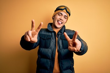 Wall Mural - Young brazilian skier man wearing snow sportswear and ski goggles over yellow background smiling with tongue out showing fingers of both hands doing victory sign. Number two.