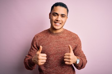 Young brazilian man wearing casual sweater standing over isolated pink background success sign doing positive gesture with hand, thumbs up smiling and happy. Cheerful expression and winner gesture.