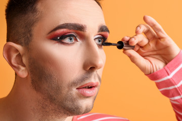 Wall Mural - Young transgender woman applying mascara onto eyelashes against color background