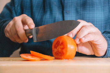 Man wearing a blue shirt slicing a tomato on a wooden board. Cooking concept