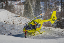 A Yellow Rescue Helicopter Is Parked In The Middle Of The Ski Slope On A Patch Of Snow. Concept Of Helicopter Rescue On Snow Or Ski Slope. Visible Pilots And Other People Around The Helicopter.