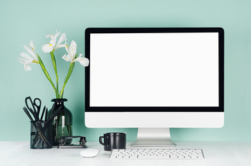 Minimalistic office interior with blank computer monitor, black stationery, books, white flowers, keyboard, coffee cup in green mint menthe interior on white wood table.