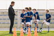 Young coach explaining training rules to children school soccer team. Kids in football team listening to coach. Boys on soccer practice. Physical education outdoor class