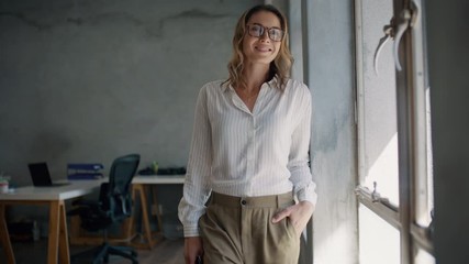 Wall Mural - Businesswoman standing by a window looking outside and smiling at camera. Female entrepreneur in formalwear looking at camera and smiling.
