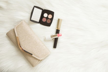 Various Decorative Makeup Cosmetics With Shiny, Golden Clutch Bag On White Fluffy Fur Blanket Background. Different Beauty Essentials For Women. Empty Place For Text. Top Down View.