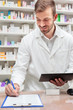 © Ivan - Handsome young male pharmacist holding a tablet and filling out paperwork on a clipboard, working in a pharmacy. Healthcare and medicine concept.