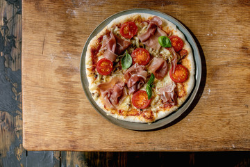 Wall Mural - Fresh baking homemade pizza napolitana with prosciutto ham, cheese, tomatoes, basil on plate over wooden table background. Home baking or delivered fast food. Flat lay, space.