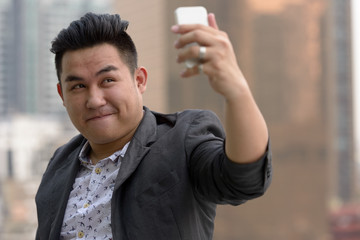 Wall Mural - Young overweight Asian businessman taking selfie against view of the city