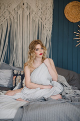 Portrait of a blonde girl sitting on a bed in a white shirt flung down from her shoulders. Horizontal photography