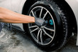 Auto wash service. Cropped close up image of male hands in black protective gloves, cleaning alloy wheels rims of luxury car with a special brush for cast wheels in a vehicle detailing workshop