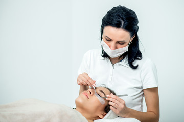 Wall Mural - Cosmetologist is doing eyelash extension procedure of patient on white background, copy space.