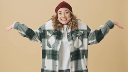 Canvas Print - Confused pretty woman in knit hat and shirt covering her ears and shrugging her shoulders while looking at the camera over beige background