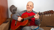 Man Plays The Classical Guitar Next To A Gray Cat On A White Background.