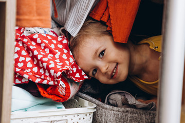 Portrait of happy smiling girl at home. Child playing with parents at home hiding in closet with clothes