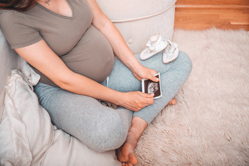 Pregnant woman holding ultrasound image. Concept of pregnancy, health care, gynecology, medicine. Young mother waiting of the baby. Close-up, copy space, indoors.