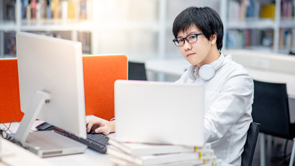 Wall Mural - Young Asian man university student using computer and laptop for educational research in college library. Male researcher finding information on internet platform. Online study or E-learning concept