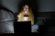 © Westend61 - Young woman sitting on the couch at home eating while using laptop