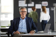 © Westend61 - Portrait of businessman at desk in office with colleagues in background