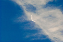 A Slim Sliver Of The Silvery Moon Set On An Expanse Of A Partly Cloudy Blue Sky.