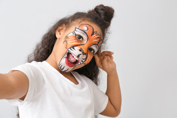 Wall Mural - Funny African-American girl with face painting taking selfie on light background