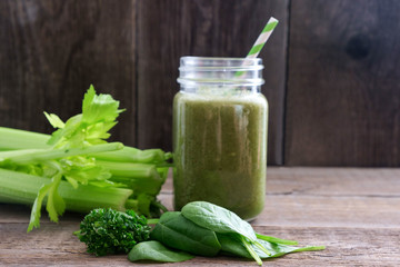 Wall Mural - A glass with a green smoothie and a bamboo cocktail straw is standing on a wooden board. Nearby are bunches of celery and spinach and parsley leaves. Useful snack. Vegan, vegetarian drink. Copy space