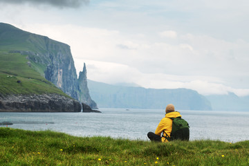 Wall Mural - Tourist with backpack in yellow jacket looks at Witches Finger cliffs from Trollkonufingur viewpoint. Vagar island, Faroe Islands, Denmark.