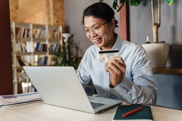 Image of joyful handsome asian man using laptop and holding credit card