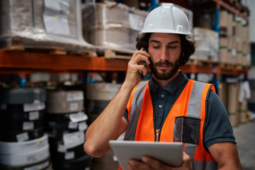 Focused warehouse manager tracking supply order details using digital tablet while in conversation over mobile phone in warehouse