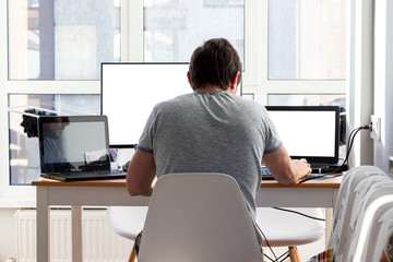 A man in a gray t-shirt is sitting at a workplace with two laptops and a monitor near the window. Remote work from home in quarantine. Back view